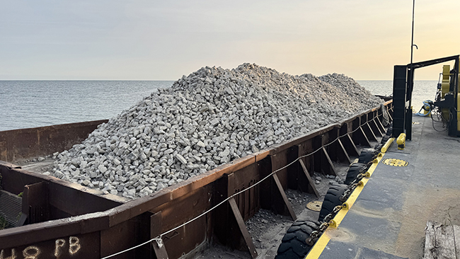 A close-up photo of a barge filled with mounds of cobble. The barge is alongside another barge that the photographer is standing on. The edge of the photographer’s barge is lined with tires.